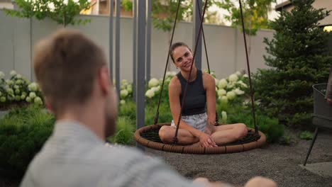 Over-the-shoulder-a-happy-brunette-girl-swings-on-a-rope-swing-and-sings-a-song-together-with-her-boyfriend-who-plays-the-guitar-in-the-backyard-of-a-country-house