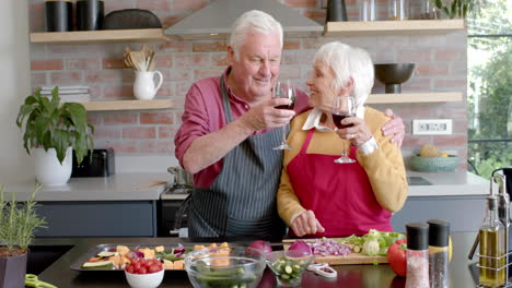 Feliz-Pareja-Caucásica-Mayor-Cocinando-La-Cena-Bebiendo-Vino-Tinto-En-La-Cocina-En-Casa,-Cámara-Lenta