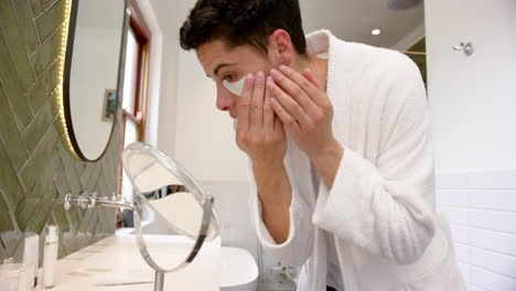 focused biracial man applying under eye mask in bathroom looking in mirror, slow motion