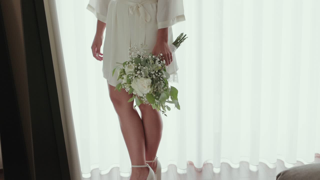 Close up of woman in white heels holding a floral bouquet near a bright curtain backdrop