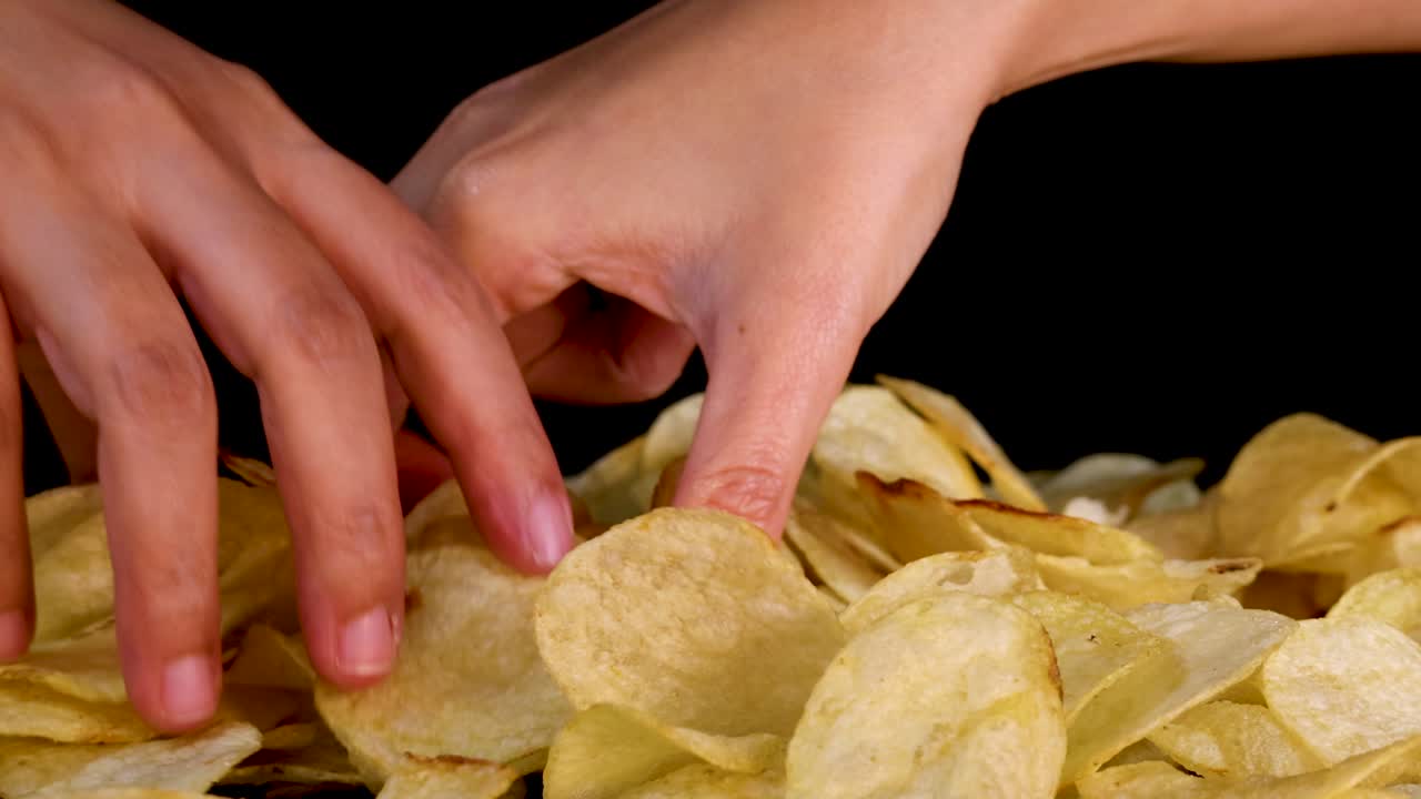 Human hand selects potato chips from pile, close-up, dramatic lighting, black background, slow movement