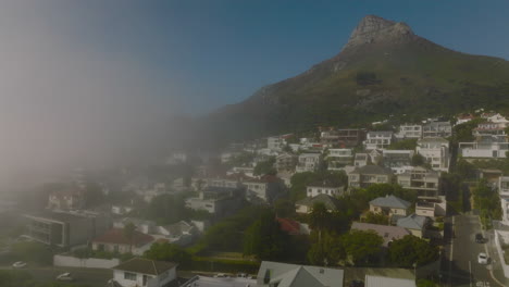 Fly-above-houses-in-residential-borough-of-touristic-destination.-High-rocky-peak-ahead.-Morning-fog-rising-from-sea.-Cape-Town,-South-Africa