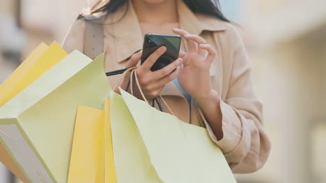 Una-Joven-Asiática-Feliz-Y-Sonriente-Con-Ropa-Elegante-Y-Moderna-Y-Bolsas-De-Compras-De-Colores-Brillantes-Está-Usando-Su-Teléfono-Inteligente-Mientras-Camina-Por-El-Centro-Comercial