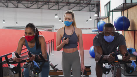 Entrenadora-De-Fitness-Femenina-Caucásica-Con-Mascarilla-En-El-Gimnasio-Parada-Junto-A-Bicicletas-Estáticas