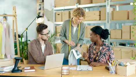 Caucasian-young-caucasian-woman-designer-showing-fabric-to-her-coworkers-who-are-typing-on-laptop-and-writing-notes-in-clothing-workshop