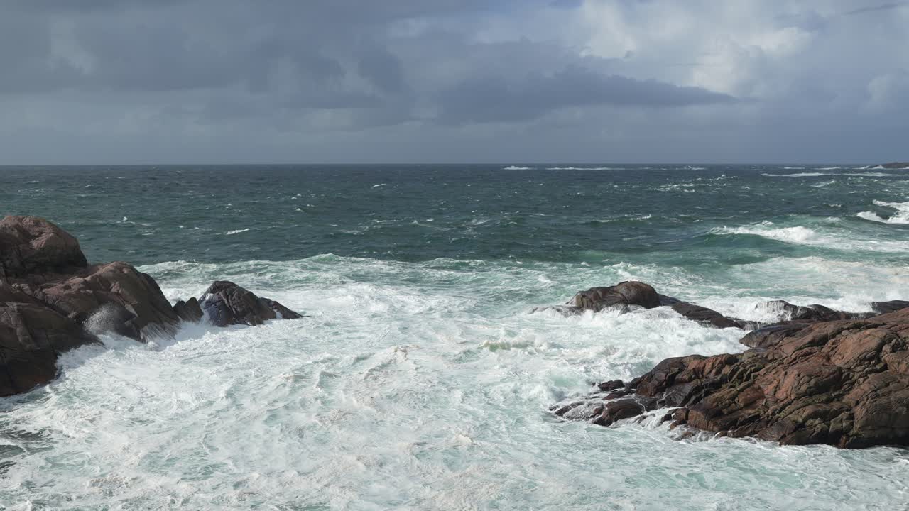 Waves crash forcefully against jagged rocks on a wild coast. The dark clouds loom overhead, hinting at a stormy atmosphere. The ocean is choppy and unpredictable