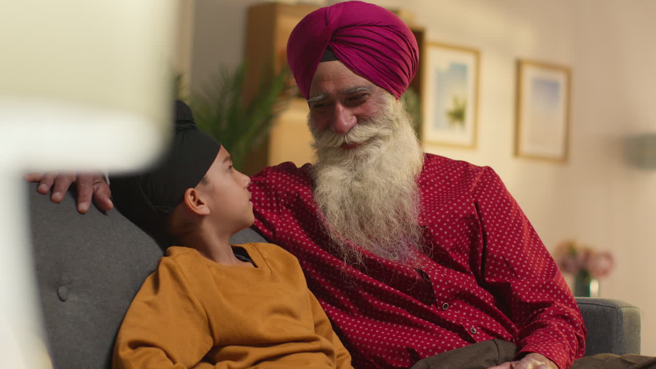 Smiling Sikh Grandfather And Grandson Wearing Turbans Sitting On Sofa At Home Talking Together