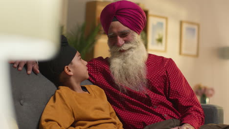 un abuelo y un nieto sikh sonrientes con turbantes sentados en el sofá en casa hablando juntos