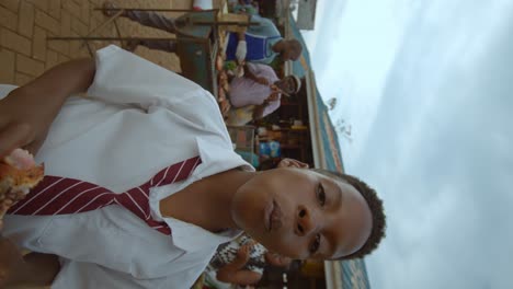 vertical - boy in uniform nodding head while eating food at african market