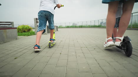 back leg view of a woman and her children riding scooters outdoors on a paved path, with iron rail by the side