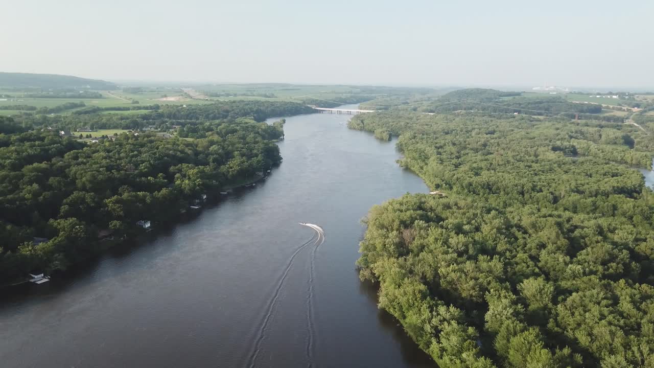 A lone speedboat travels up a winding river cutting through dense green forest, leaving a white wake behind on the calm water as hills rise in the distance.