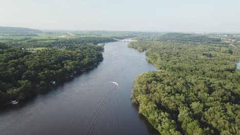 a lone speedboat travels up a winding river cutting through dense green forest, leaving a white wake behind on the calm water as hills rise in the distance.