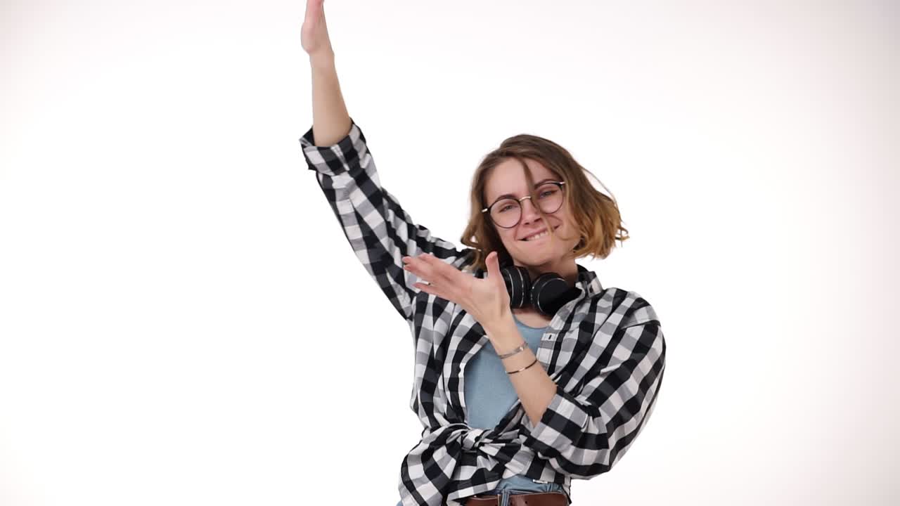 Slow motion of beautiful curly short haired girl winning a lot of money and throwing dollar bills in the air from hands isolated over white background in studio. Wearing plaid shirt, eyeglasses and headphones on neck