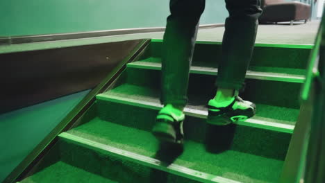 Male foot in black and white sneaker stepping up stairs under green lighting, walking towards billiard room. Carpeted stairs and metal railings add modern ambiance with a dynamic sense of movement