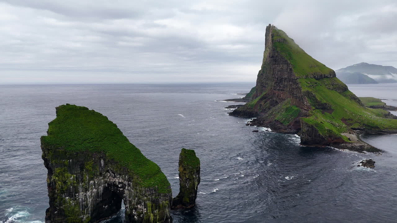 Cinematic aerial view of Drangarnir sea stacks rising dramatically from the Atlantic Ocean near Vágar, Faroe Islands, showcasing rugged cliffs, lush green slopes, and misty Nordic seascape