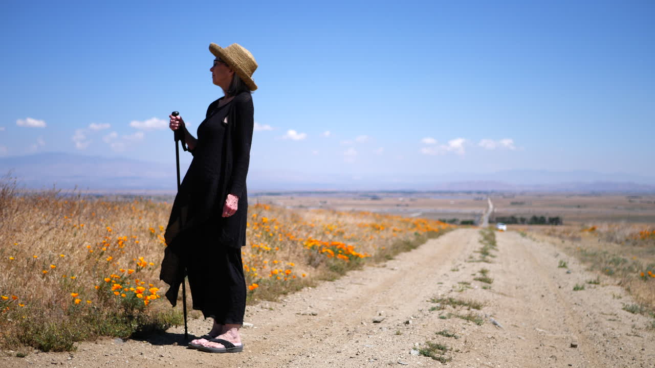 una anciana con un espectacular vestido negro y sombrero para el sol en un camino de tierra bajo un cielo azul con flores y viento soplando a cámara lenta
