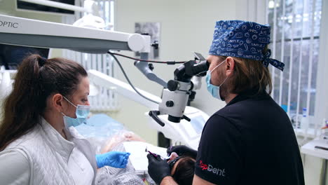 focused male dentist looks into binoculars of endodontic microscope. doctor holding tools in both hands moving them carefully in patient's mouth. nurse watching the process.