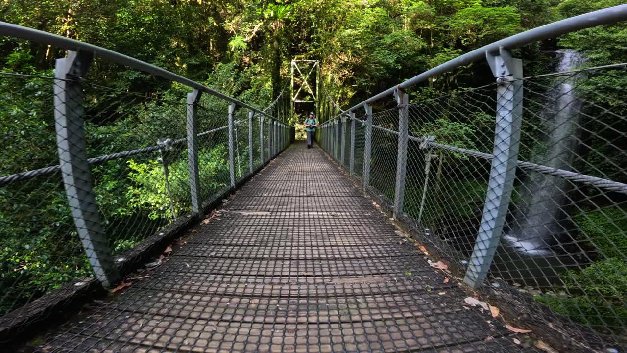 A man walks across a suspension bridge in Dorrigo National Park, surrounded by lush rainforest and cascading waterfalls