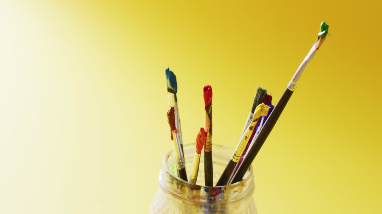 video de una colección de pinceles usados de pie en un frasco sobre un fondo amarillo con espacio de copia