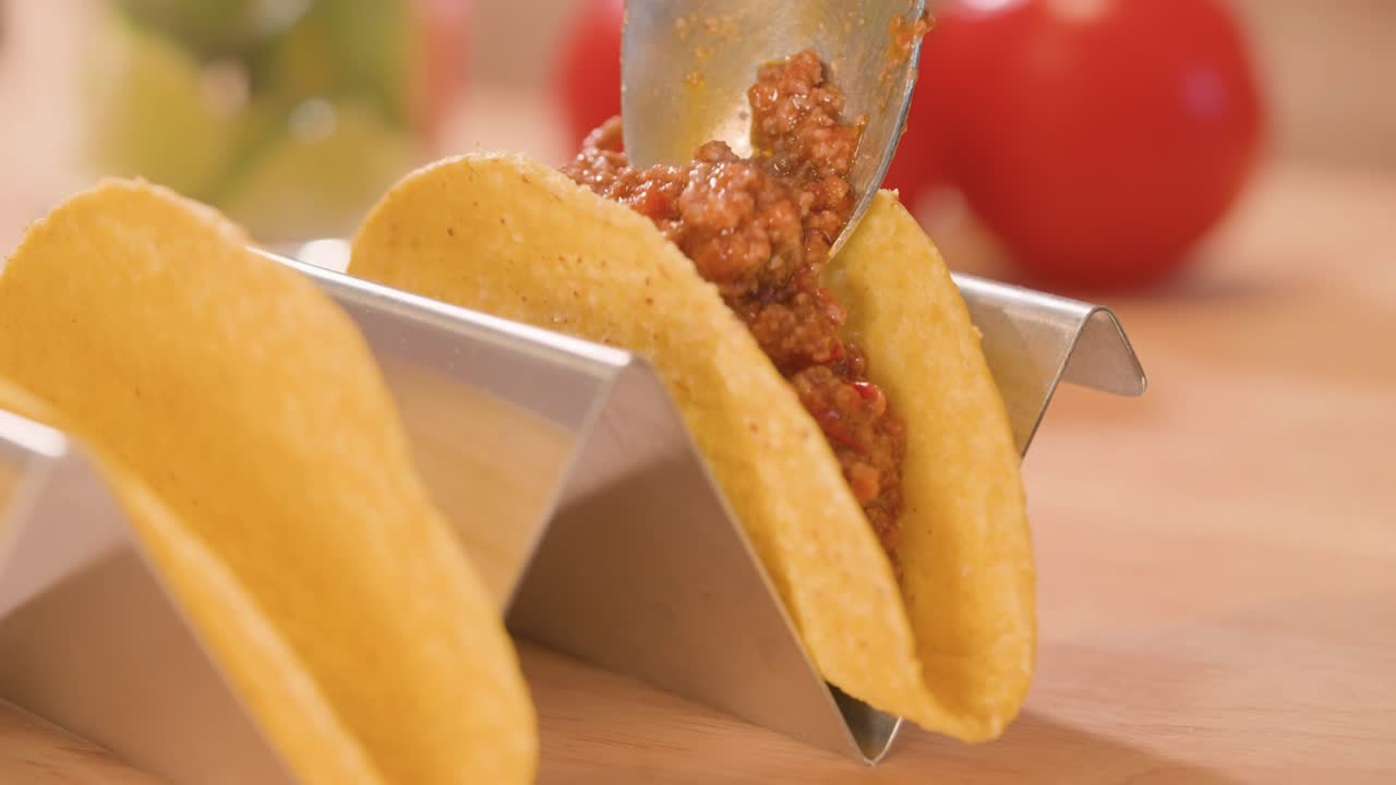 Close-up of hard corn taco shells, partially filled with seasoned meat and a spoonful being added, on a stainless steel taco stand with one empty shell.