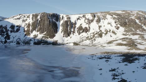 aerial drone footage flying close above a beautiful frozen loch etchachan towards a dramatic and imposing ice-covered rock face and cliff with steep snow filled gullies