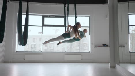 Dos-Jóvenes-Yoguis-Haciendo-Práctica-De-Yoga-Aéreo-En-Hamacas-Verdes-En-El-Gimnasio.-Hermosas-Mujeres-Trabajando-En-Clase-Realizando-Aero-Yoga.-Variación-De-La-Postura-Piramidal-Parsvottanasana.