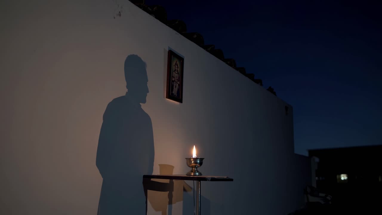 Shadow of a priest praying in front of a religious picture and a burning candle on a small table, with a white wall and a dark blue sky in the background at night