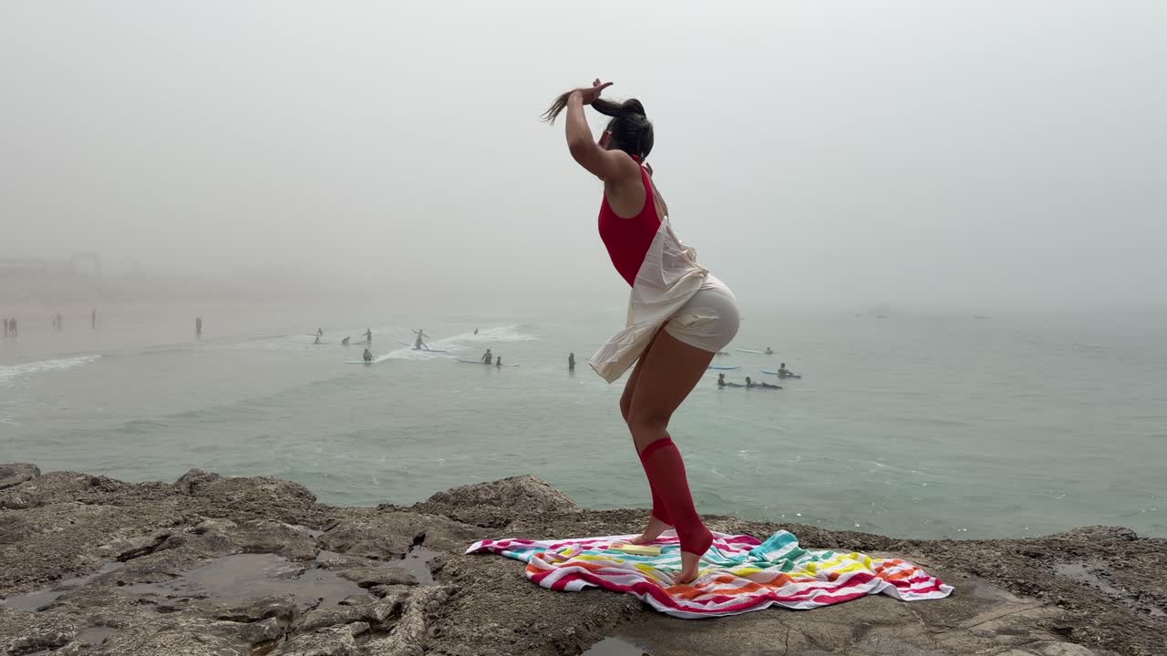 Mulher aplicando protetor solar em um penhasco de praia nebuloso com vista para surfistas