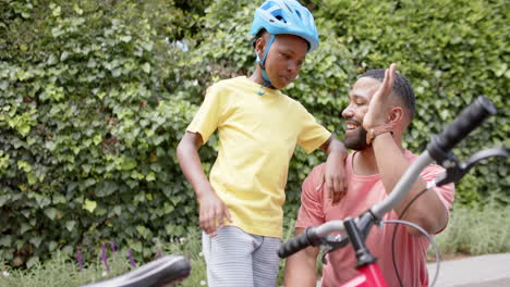 happy african american father and son with bicycle high fiving in garden, slow motion