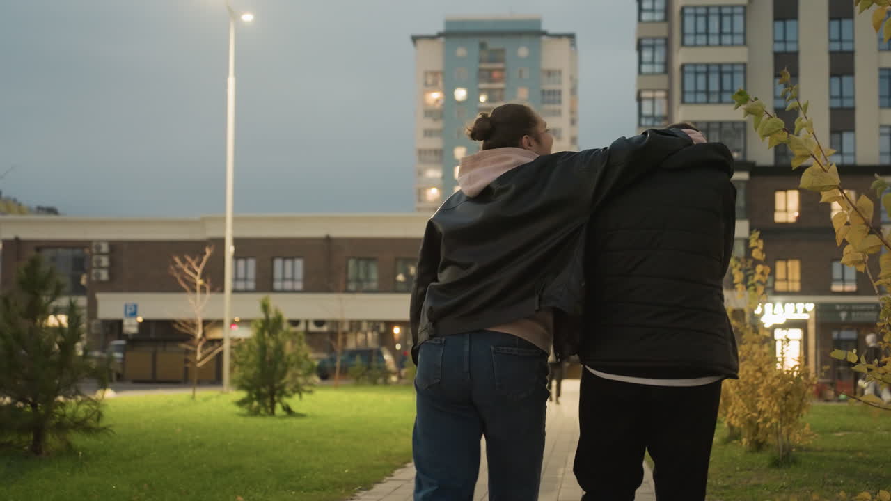 Rear view of woman playfully touching her friend head as she holds him, and he playfully rubs her head, walking together in an urban setting with buildings and lights in the background