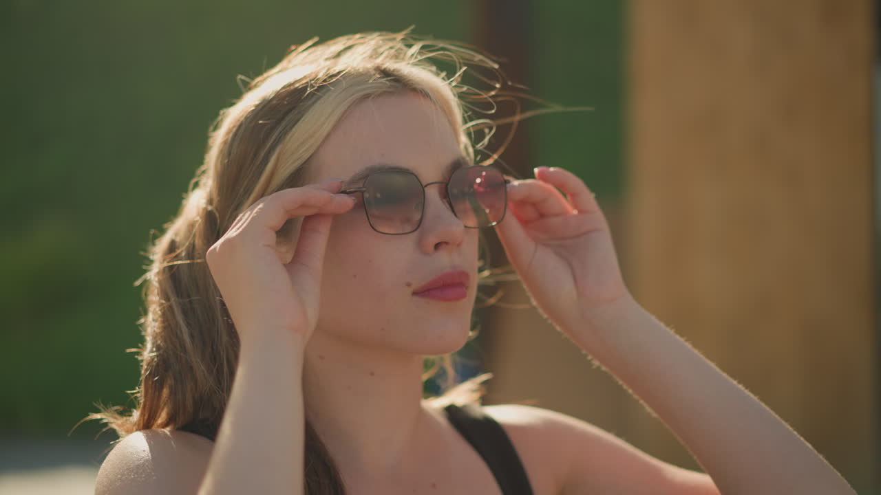 야외에 앉아있는 여자는 머리에서 선글라스를 내려놓고, 태양빛이 그녀에 반사되는 동안 자신감있는 표정으로 착용합니다. 배경에는 초록색과 목조 구조가 있습니다.
