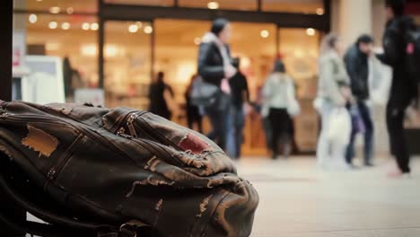 a worn-out backpack rests on the floor of a bustling shopping center as shoppers pass by, creating a vivid contrast between the still bag and the moving crowd.