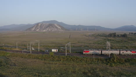 russian railway train locomotives pulling a long empty coal carriages freight through green fields with mountains in the background on the sunset, russian federation