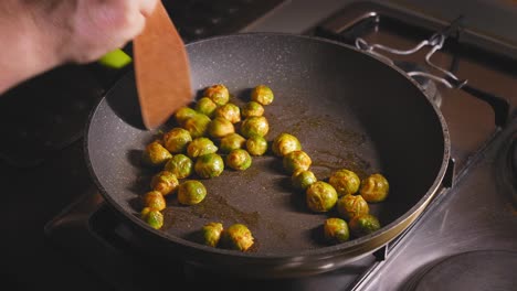 un primer plano de las coles de bruselas marinadas asadas en una placa de gas en una cocina, mientras un chef tira y remueve cuidadosamente las verduras con una espátula de madera para garantizar una cocción uniforme en una sartén caliente