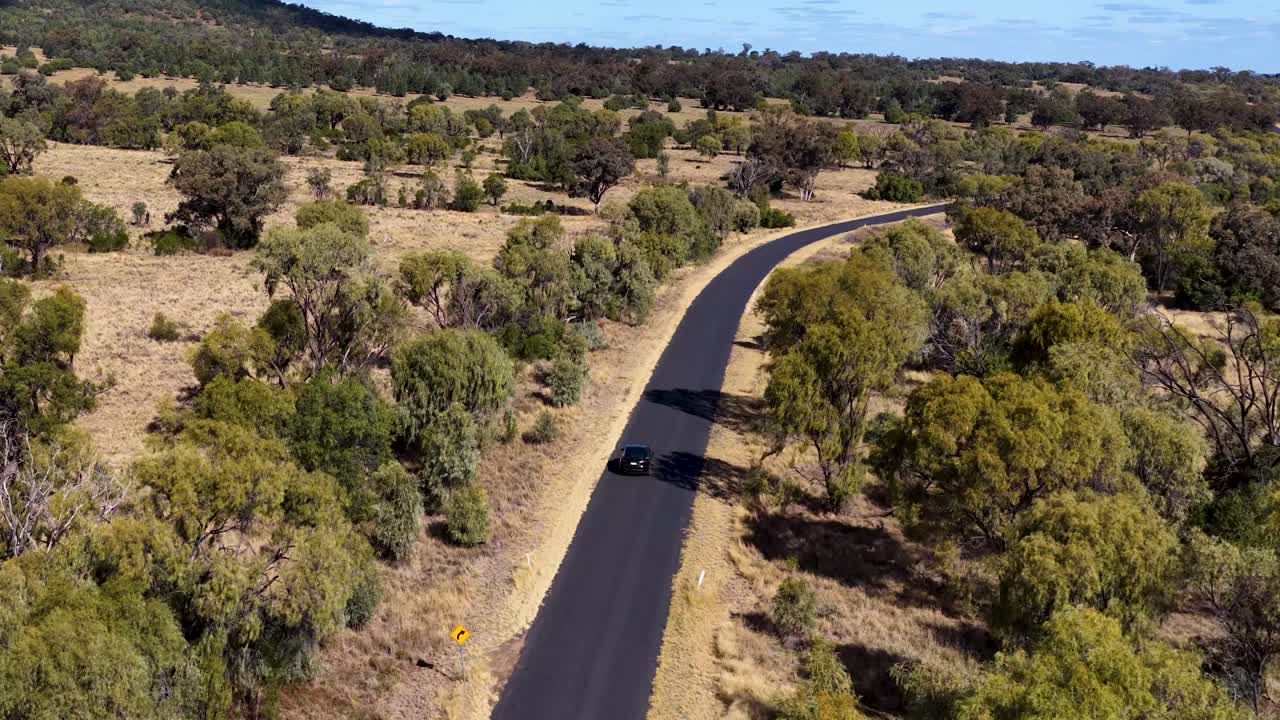 A black electric vehicle drives along a winding asphalt road through dry bushland, filmed by a smoothly tracking aerial drone in bright daylight