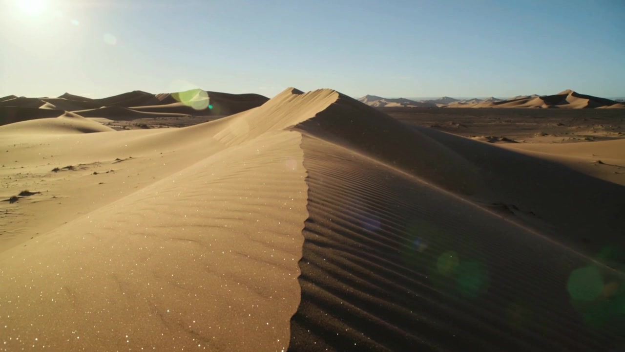La telecamera si allontana, riprendendo la cresta centrale di una duna di sabbia nel deserto, rivelando increspature e bagliori dell'obiettivo.