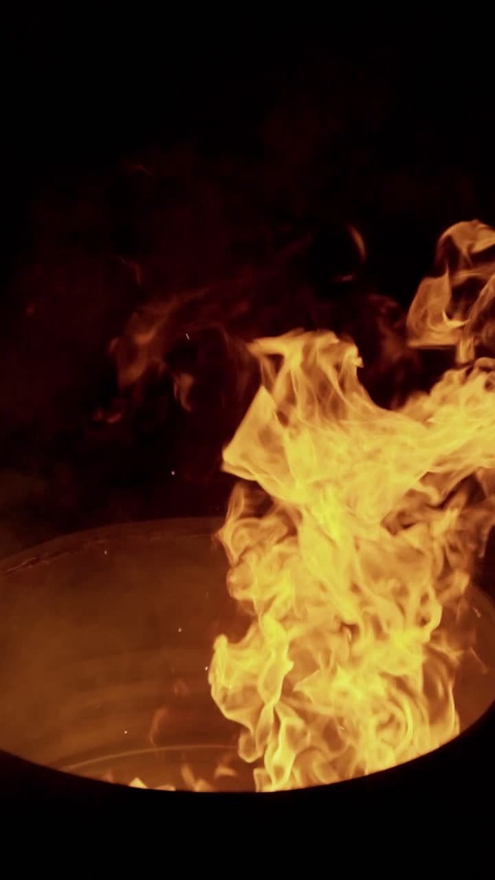Primo piano cinematografico in 4K della mano di una persona che posiziona un pezzo di legno in un falò scoppiettante da campeggio di notte.