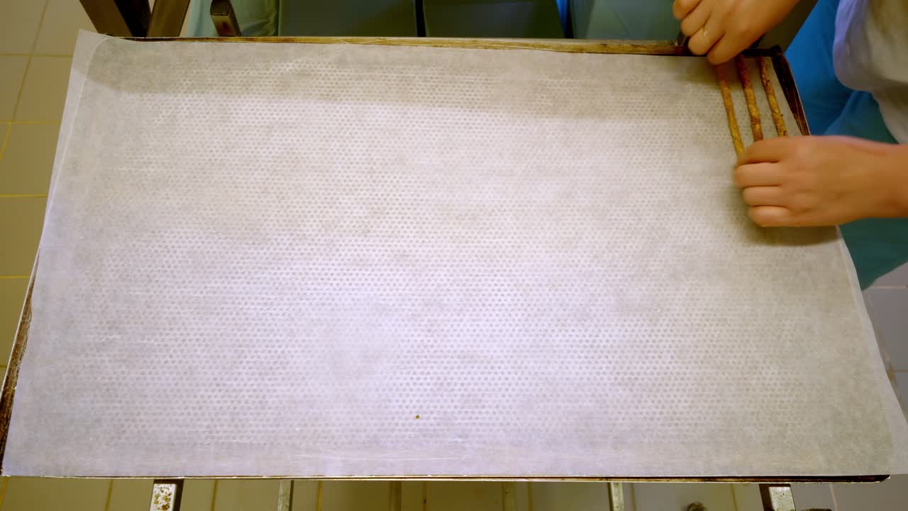 Top view of baker preparing dough strips on baking tray with parchment paper. Food production process showing pastry preparation in bakery or industrial kitchen environment