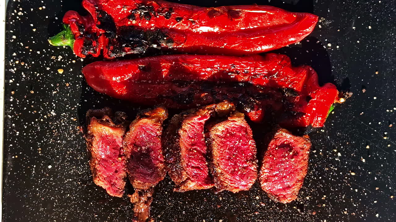An extreme close-up shot shows a perfectly grilled beef steak, sliced to reveal its juicy, rare red interior, served alongside charred red peppers on a dark plate