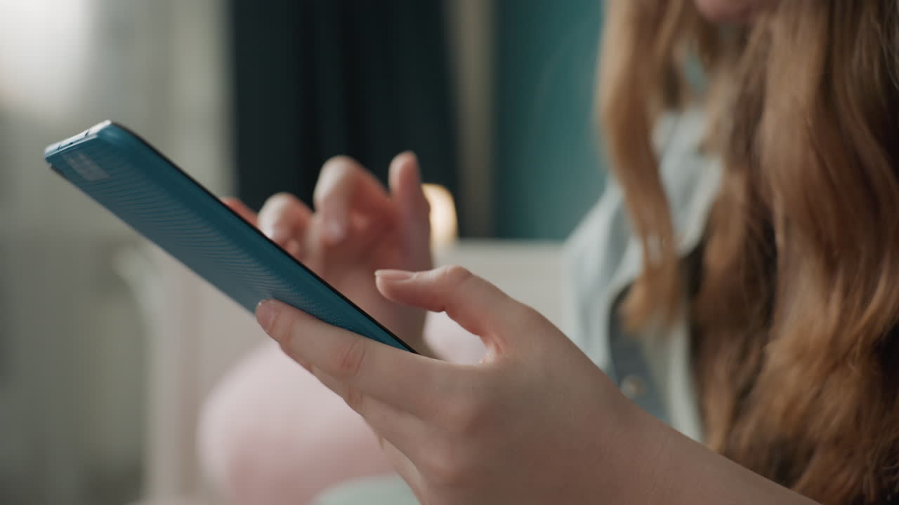Primo piano di mani caucasiche che tengono uno smartphone, studente che invia messaggi in una camera da letto accogliente, luce naturale soffusa, cover blu del telefono, polpastrello concentrato che tocca lo schermo, atmosfera di vita domestica rilassata, momento sociale spontaneo.