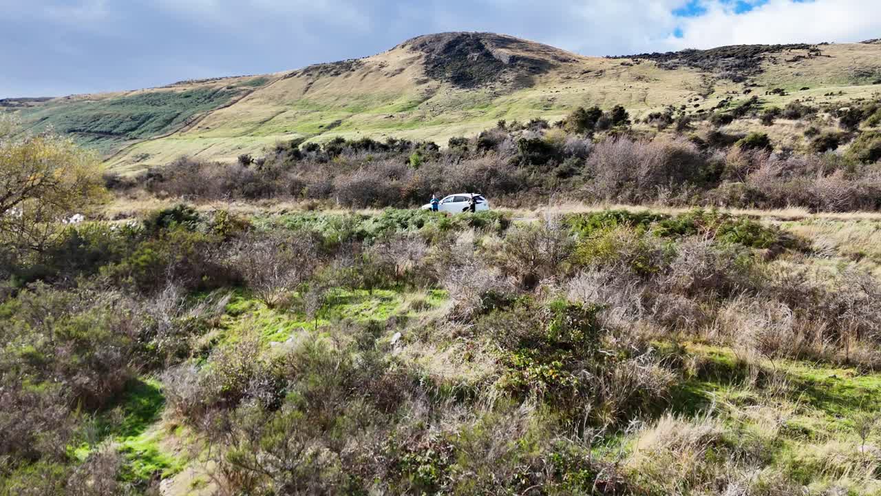 A car travels through Glenorchy's picturesque terrain, surrounded by lush vegetation and distant hills under clear skies