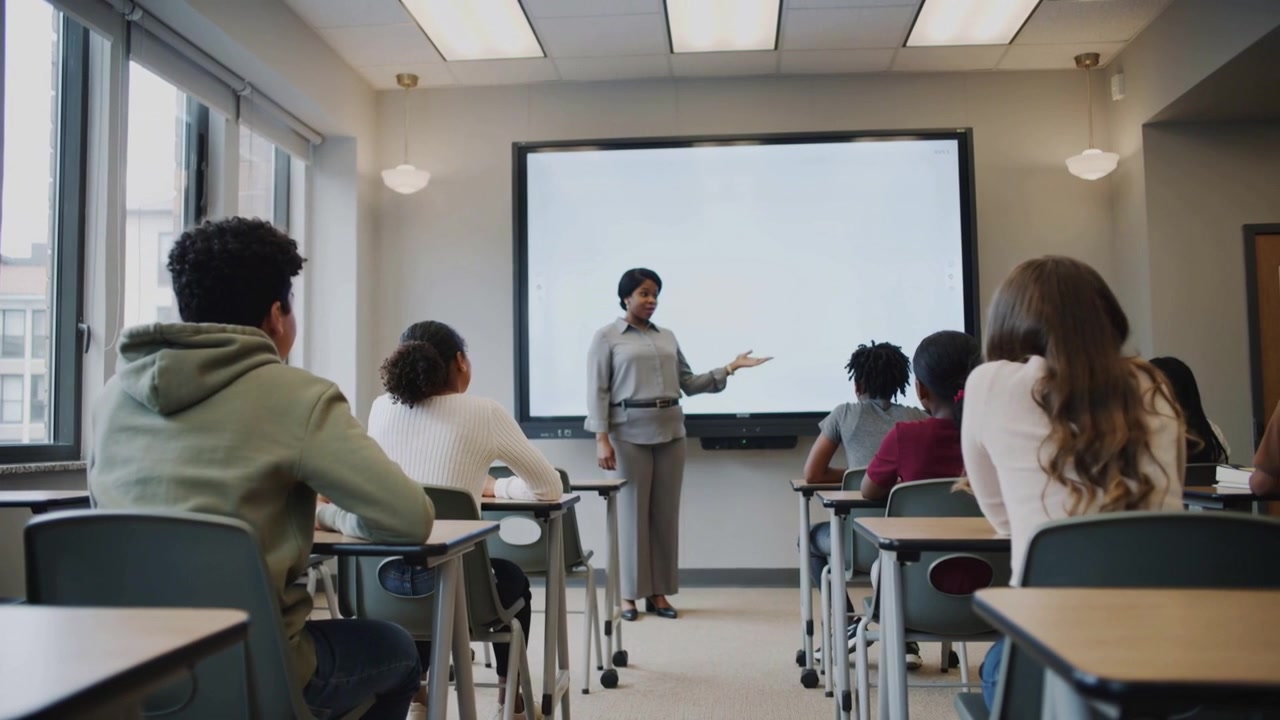 Inizio della lezione, l'insegnante in camicetta grigia gesticola davanti alla classe verso la lavagna digitale, insegnando.