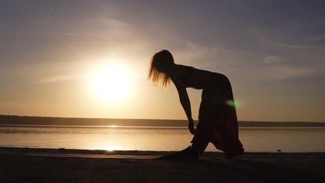 La-Silueta-De-Una-Mujer-En-Forma-Cerca-Del-Agua-Despliega-La-Estera-De-Yoga,-Preparándose-Para-Hacer-Yoga-O-Pilates.-Playa-Al-Atardecer