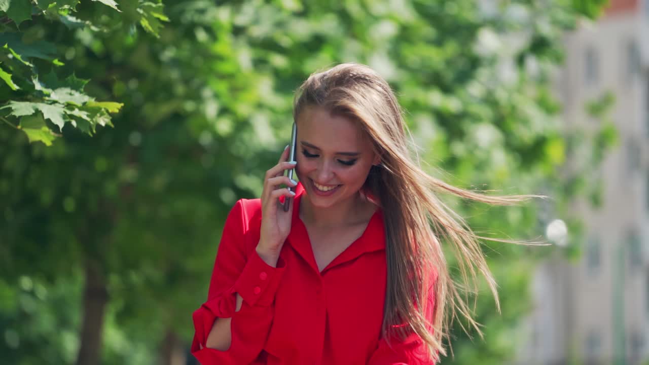 Happy woman talking on the cellphone. Attractive smiling young lady in red dress walking in the city and talks on the mobile phone. Slow motion.