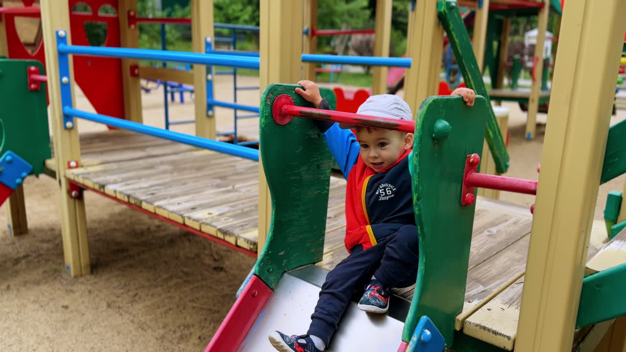 Cute baby boy goes up on the slide. Toddler sits down tapping his feet actively and slides down.