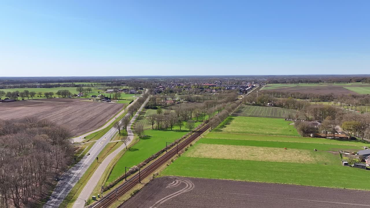 Aerial view of rural landscape with fields, trees, and distant settlement district in sunlight.