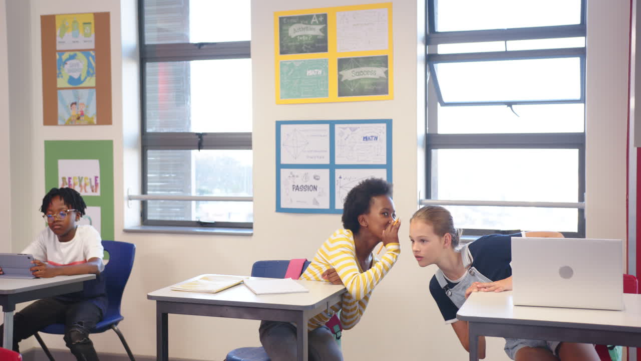 In school, two girls whispering while boy using tablet in classroom