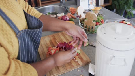 Midsection-of-african-american-woman-in-apron-composting-vegetable-waste-in-kitchen,-slow-motion