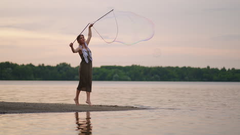 a young female artist shows a soap bubble show blowing up huge soap bubbles on the shore of a lake at sunset. show a beautiful show of soap bubbles in slow motion