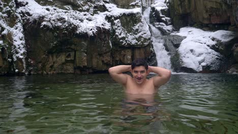 handsome man standing in subzero cold water by the beautiful waterfall in snowy mountains during winter day - static shot, slow motion